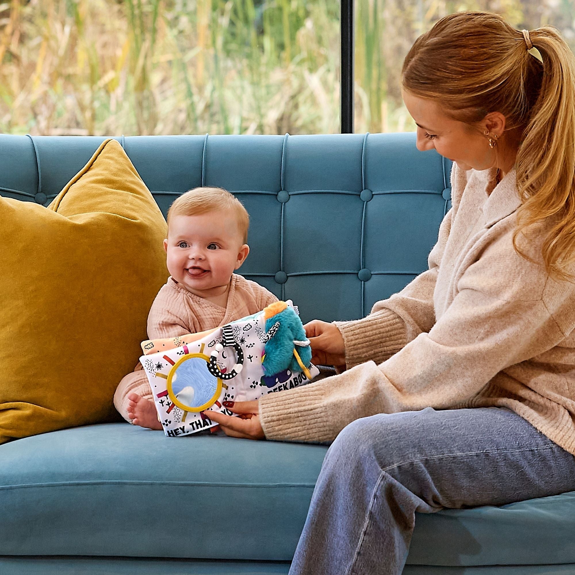 A smiling baby sits on a blue couch with a yellow pillow, holding the Things with Wings Peek a Boo Book, as an adult woman with a ponytail in a beige sweater and jeans sits beside them.