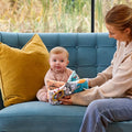 A baby sits on a blue sofa next to a yellow pillow, holding the Things with Wings Peek a Boo Book. An adult in a beige sweater sits beside the baby, offering support.