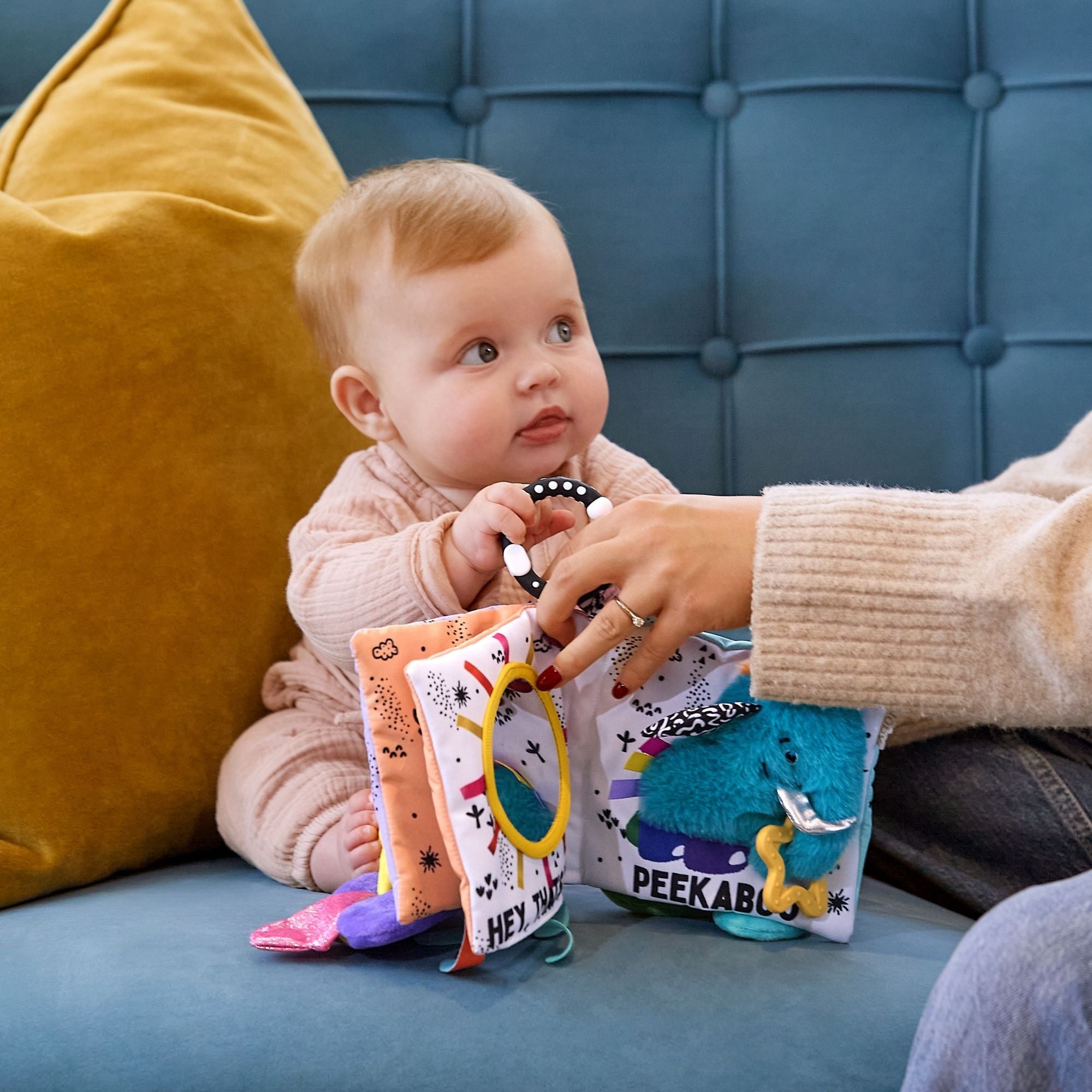 A baby sits on a blue couch, exploring the Things with Wings Peek a Boo Book—a colorful cloth book with textured pages and “PEEKABU” wording—while an adult helps. A yellow pillow is in the background.