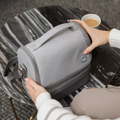 A person in a striped sweater holds a light gray insulated lunch bag on a black and white marble table, with coffee and the Nuby UK RapidCool Bottle Maker and Portable UV Steriliser Bundle nearby.