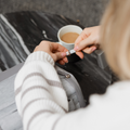 A person in a white and striped sweater plugs a USB cable into the Nuby UK RapidCool Bottle Maker and Portable UV Steriliser Bundle on a marble table, with a cup of coffee nearby.