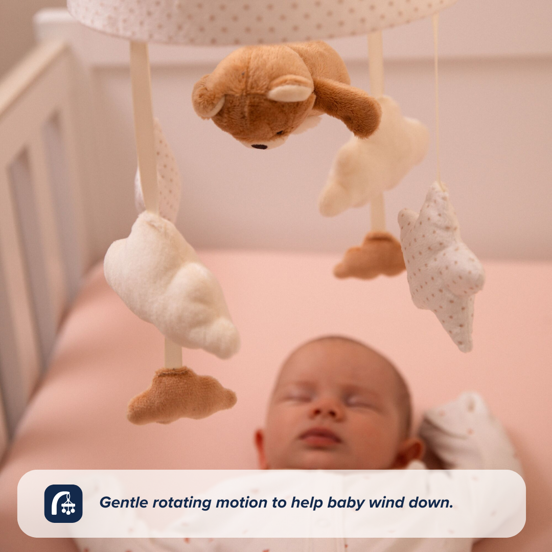 A sleeping baby lies in a crib under the Nuby UK Biscuit Bear Musical Cot Mobile, featuring plush clouds and a bear, with the caption: Gentle rotating motion and calming lullabies to help baby wind down.