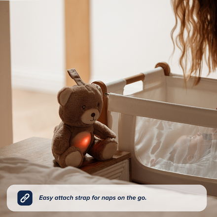 The Nuby UK Biscuit Bear Sleep Aid with Cry Sensor, featuring a glowing red heart, soothing sounds, and an easy-attach strap, sits next to a white bassinet. A womans hair is visible in the background.