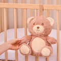 A hand touches the Biscuit Bear Baby Sleep Aid with Cry Sensor, a soft brown and beige plush hanging from a wooden crib with white bedding.