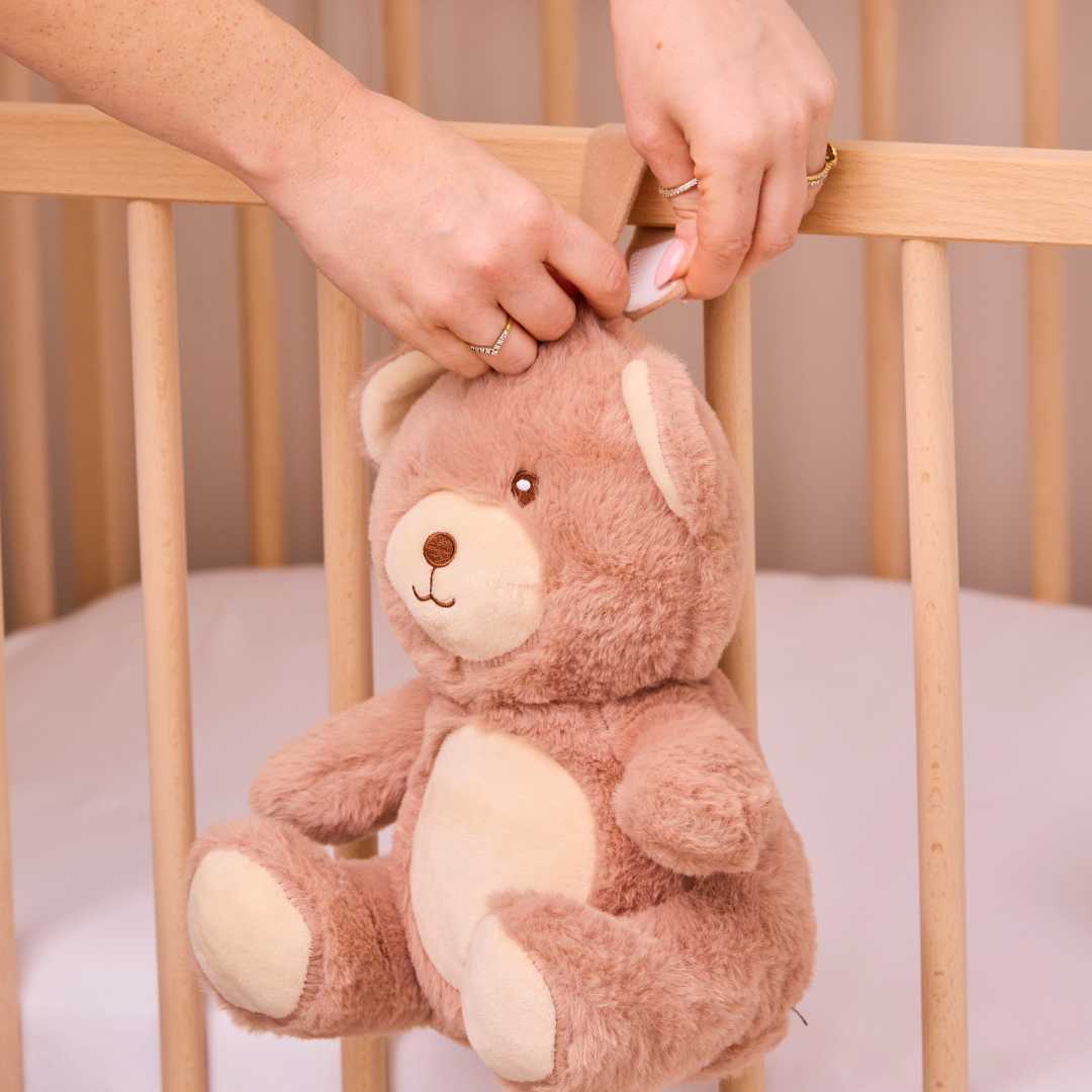 A person attaches the Biscuit Bear Baby Sleep Aid with Cry Sensor, a brown teddy bear, to a wooden crib rail with white bedding visible inside.