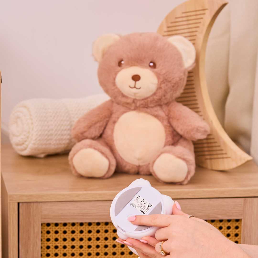 A plush teddy bear sits on a wooden cabinet beside a rolled white blanket and curved comb, as a hand holds the Biscuit Bear Baby Sleep Aid with Cry Sensor, its label visible in the foreground.