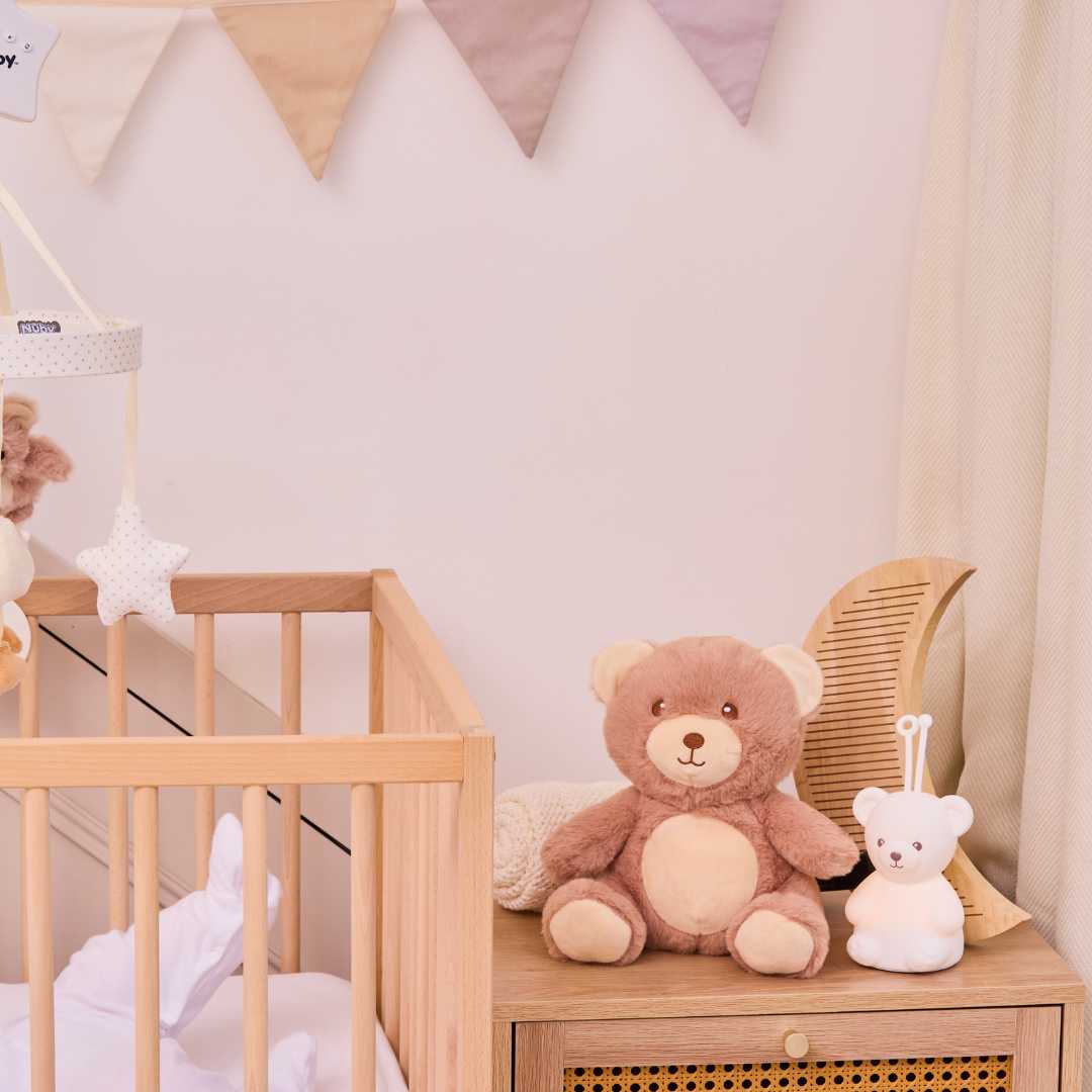 A cozy nursery with a wooden crib, brown teddy bear, small white bear, wicker basket on the dresser, and the Biscuit Bear Baby Sleep Aid with Cry Sensor glowing softly as neutral bunting hangs above the crib.