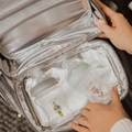 A person opens a gray Nuby UK bag with a white interior holding two animal-illustrated bottles from the RapidCool Bottle Maker and Portable UV Steriliser Bundle, placing the bottles side by side next to the steriliser.