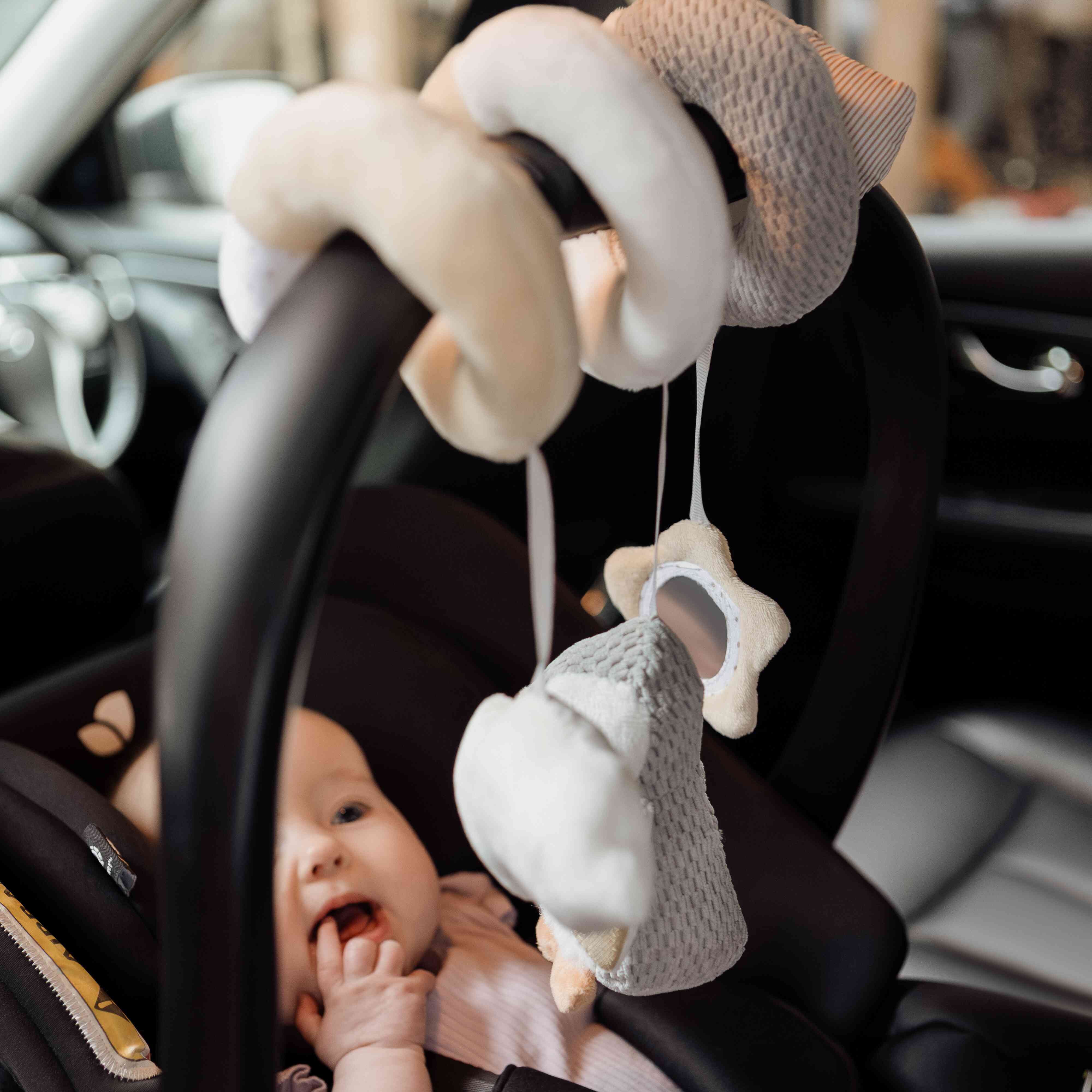 A baby sits in a car seat, gazing curiously at the Nuby Penguin Activity Spiral Travel Pram Toy hanging from the handle, touching their mouth while reaching toward the engaging toy.