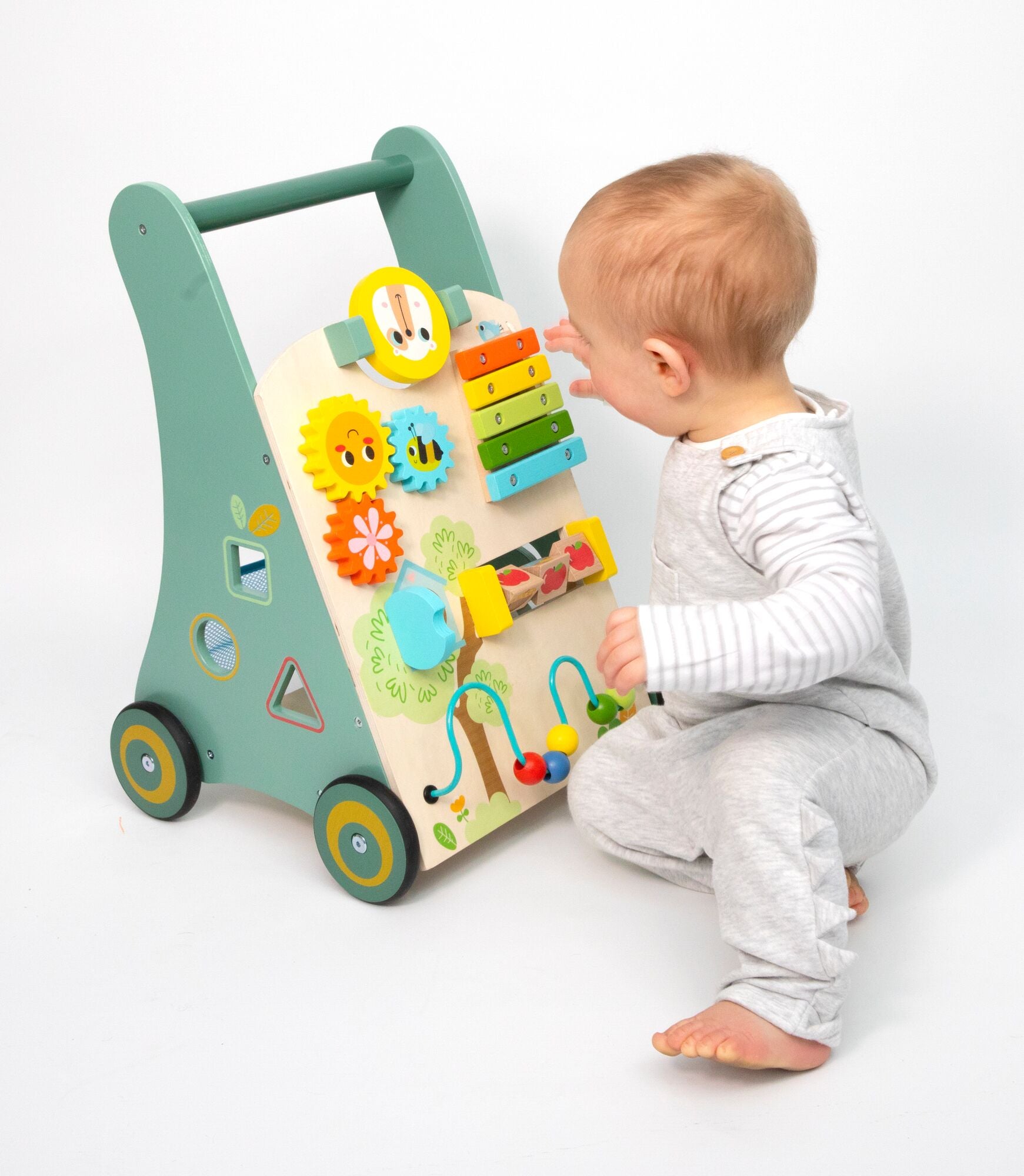 A baby in light gray clothes sits on the floor, playing with the Nuby Wooden Baby Walker. This colorful multi-activity toy features gears and a xylophone, serving as both a walking aid and interactive play center on a white background.