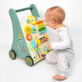 A baby in light gray clothes sits on the floor, playing with the Nuby Wooden Baby Walker. This colorful multi-activity toy features gears and a xylophone, serving as both a walking aid and interactive play center on a white background.