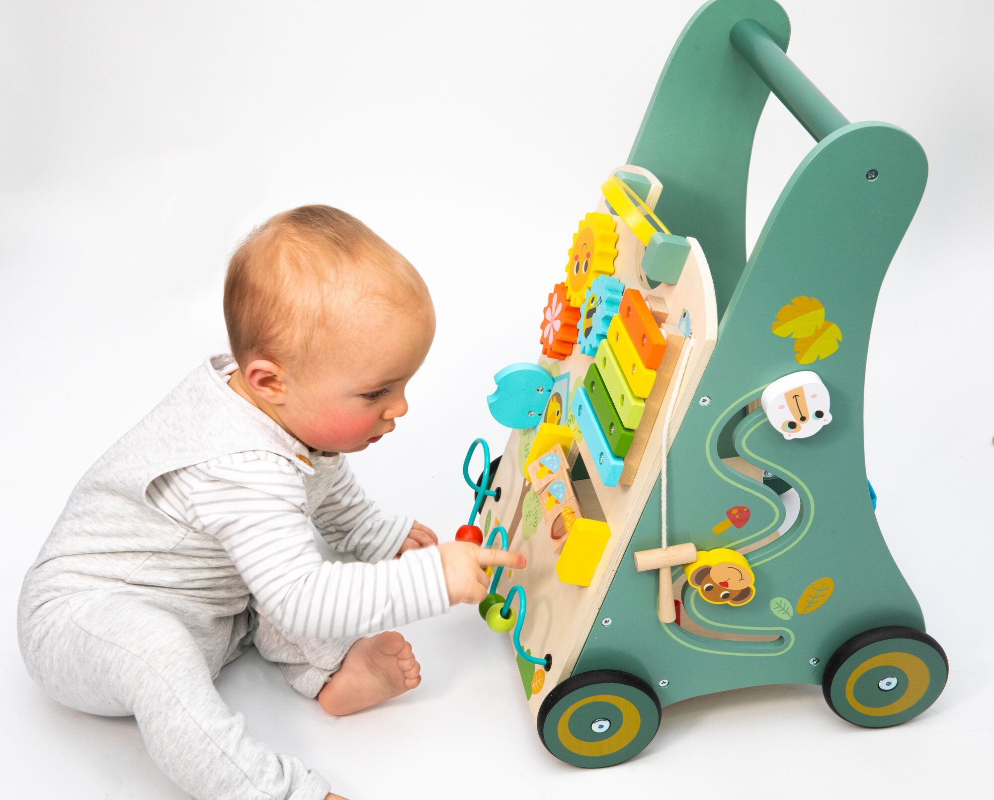 A baby in a white outfit sits on the floor, playing with the Nuby Wooden Baby Walker featuring colorful beads, gears, and animal shapes on a white background.