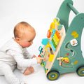 A baby in a white outfit sits on the floor, playing with the Nuby Wooden Baby Walker featuring colorful beads, gears, and animal shapes on a white background.