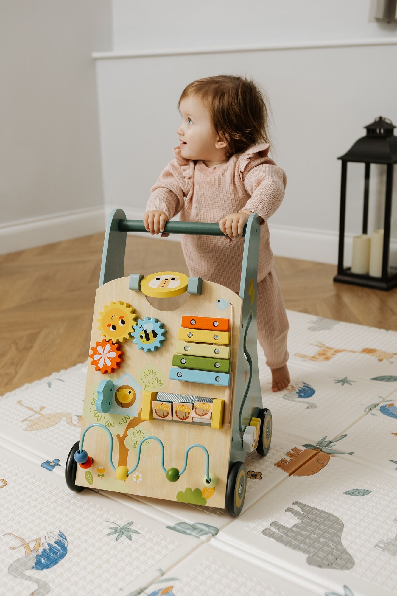 A toddler in a pink outfit stands holding the Nuby Wooden Baby Walker, featuring colorful gears, a xylophone, and beads, on an animal-themed play mat in a cozy room.