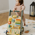 A toddler in a pink outfit stands holding the Nuby Wooden Baby Walker, featuring colorful gears, a xylophone, and beads, on an animal-themed play mat in a cozy room.