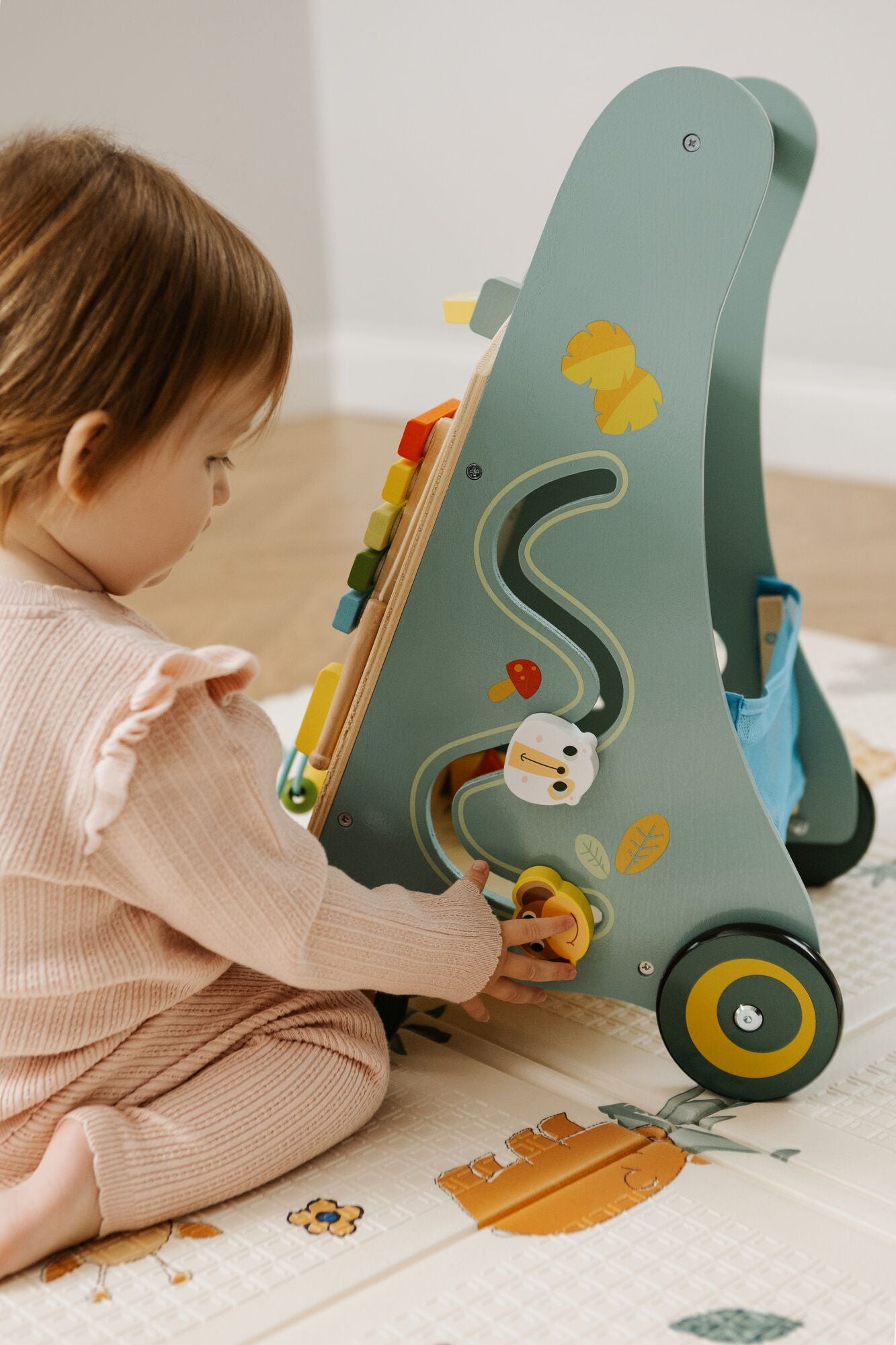 A toddler in a pink outfit sits on a play mat, playing with the Nuby Wooden Baby Walker, which features colorful beads, a sliding toy, and printed animal designs.