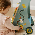 A toddler in a pink outfit sits on a play mat, playing with the Nuby Wooden Baby Walker, which features colorful beads, a sliding toy, and printed animal designs.