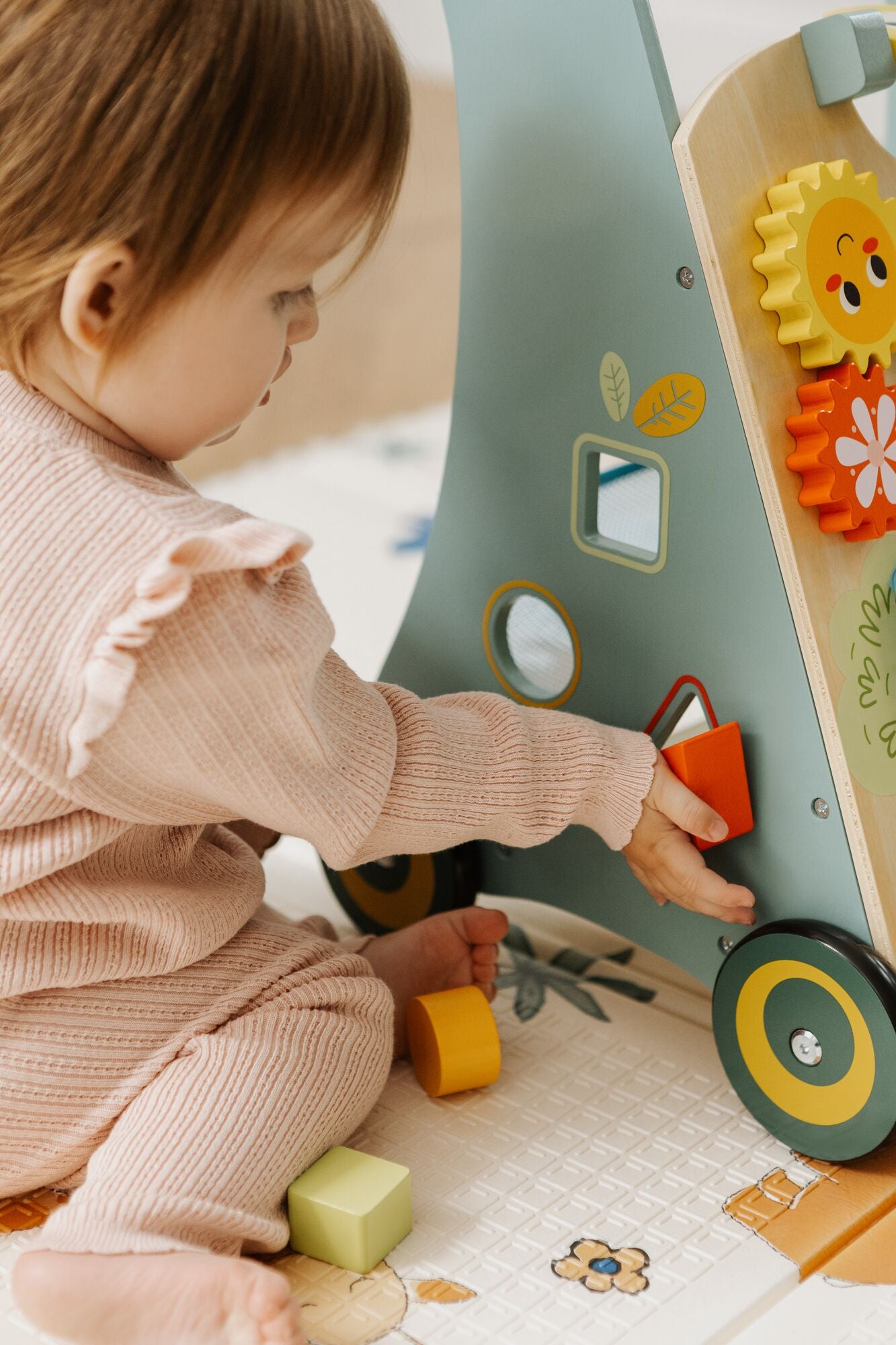 A young child in a pink outfit sits on the floor, playing with the Nuby Wooden Baby Walker, which features colorful gears, mirrors, and shape-sorting blocks.
