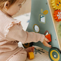A young child in a pink outfit sits on the floor, playing with the Nuby Wooden Baby Walker, which features colorful gears, mirrors, and shape-sorting blocks.