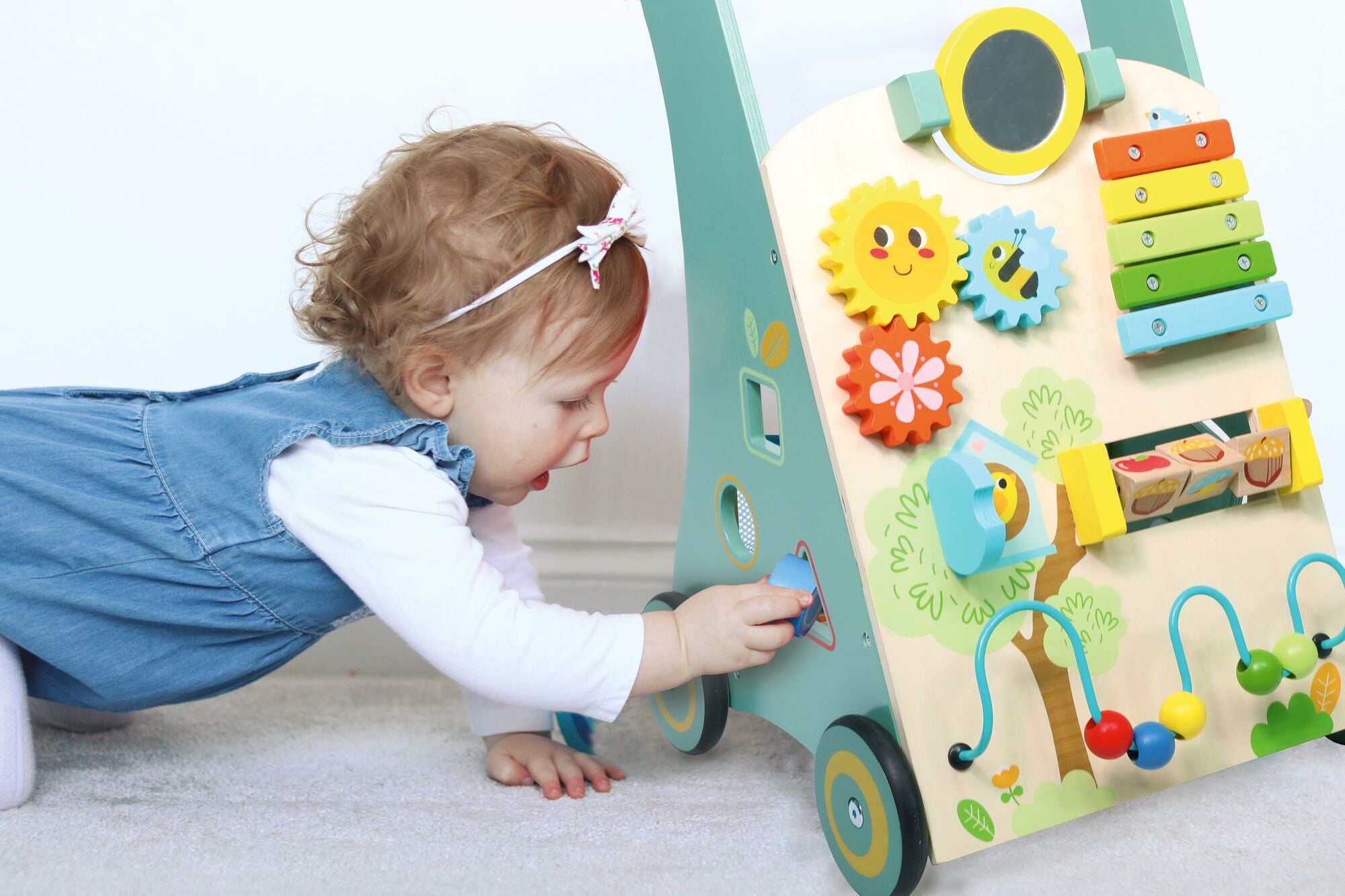 A toddler with curly hair and a headband, wearing a blue dress, plays on the floor with the Nuby Wooden Baby Walker, which features colorful gears, a xylophone, and other interactive elements.