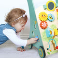 A toddler with curly hair and a headband, wearing a blue dress, plays on the floor with the Nuby Wooden Baby Walker, which features colorful gears, a xylophone, and other interactive elements.