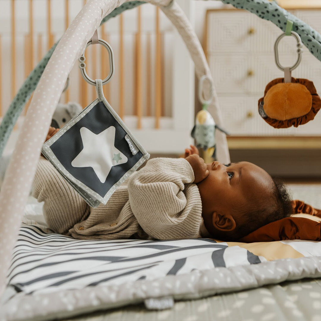 A baby relaxes on Nuby UK’s Animal Adventures Nursery Bundle playmat beneath a soft activity gym, reaching for hanging toys like a star and animal in a cozy nursery.