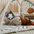 A baby relaxes on Nuby UK’s Animal Adventures Nursery Bundle playmat beneath a soft activity gym, reaching for hanging toys like a star and animal in a cozy nursery.