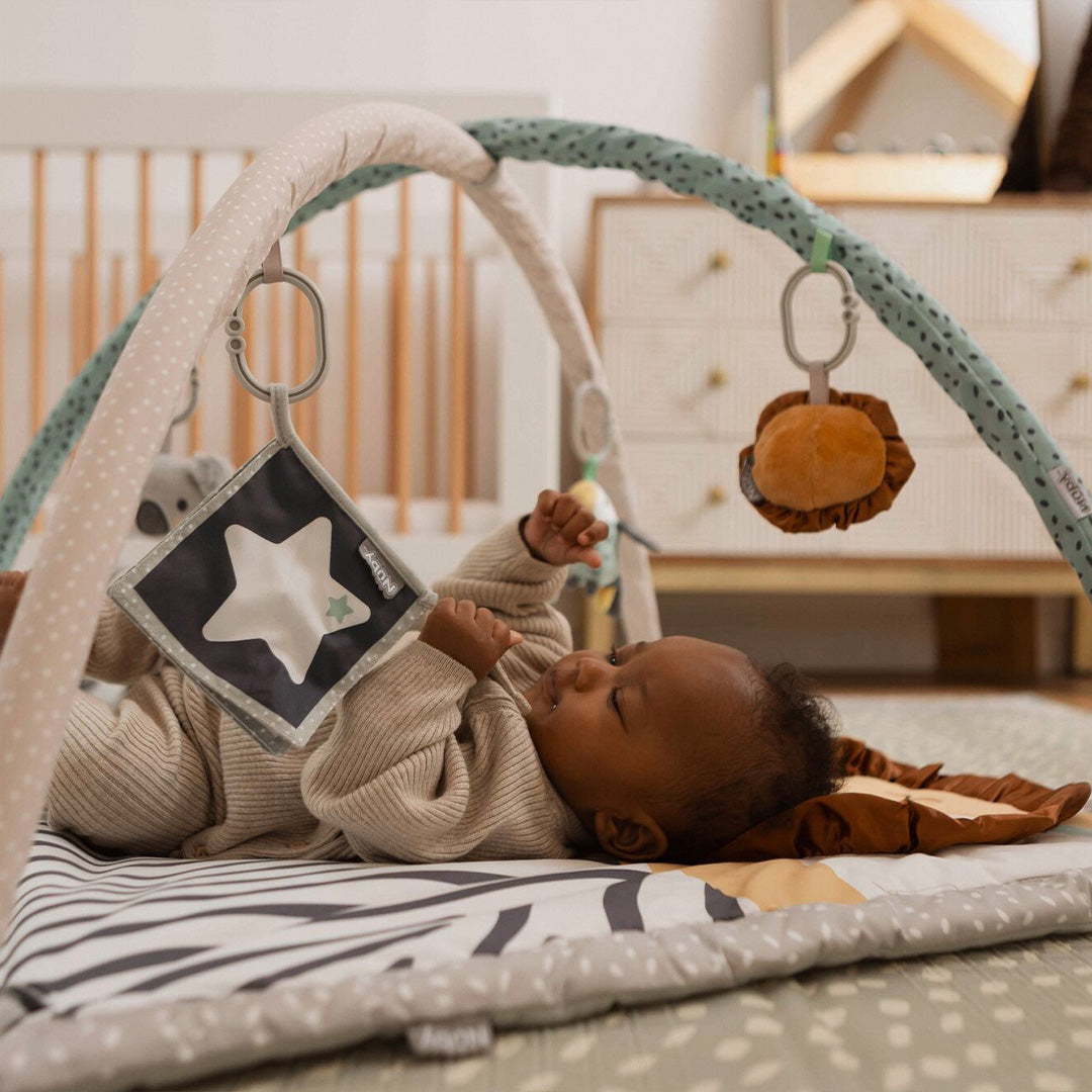 A baby rests on the Nuby Animal Adventures Baby Play Mat with Gym, enjoying hanging sensory toys like a star and plush round toy, in a cozy nursery with a crib and dresser in the background.