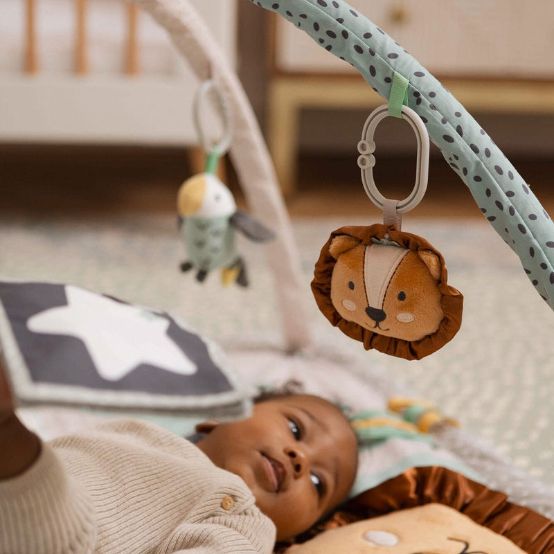 A baby in beige lies on the Nuby Animal Adventures Baby Play Mat with Gym, reaching for a plush lion toy while a sensory bird toy hangs nearby. The baby looks curious and engaged with the activity gym.