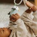 A baby in a beige ribbed outfit lies on the Nuby Animal Adventures Baby Play Mat with Gym, reaching up to hold a plush bird toy hanging above, with their feet playfully in the air.