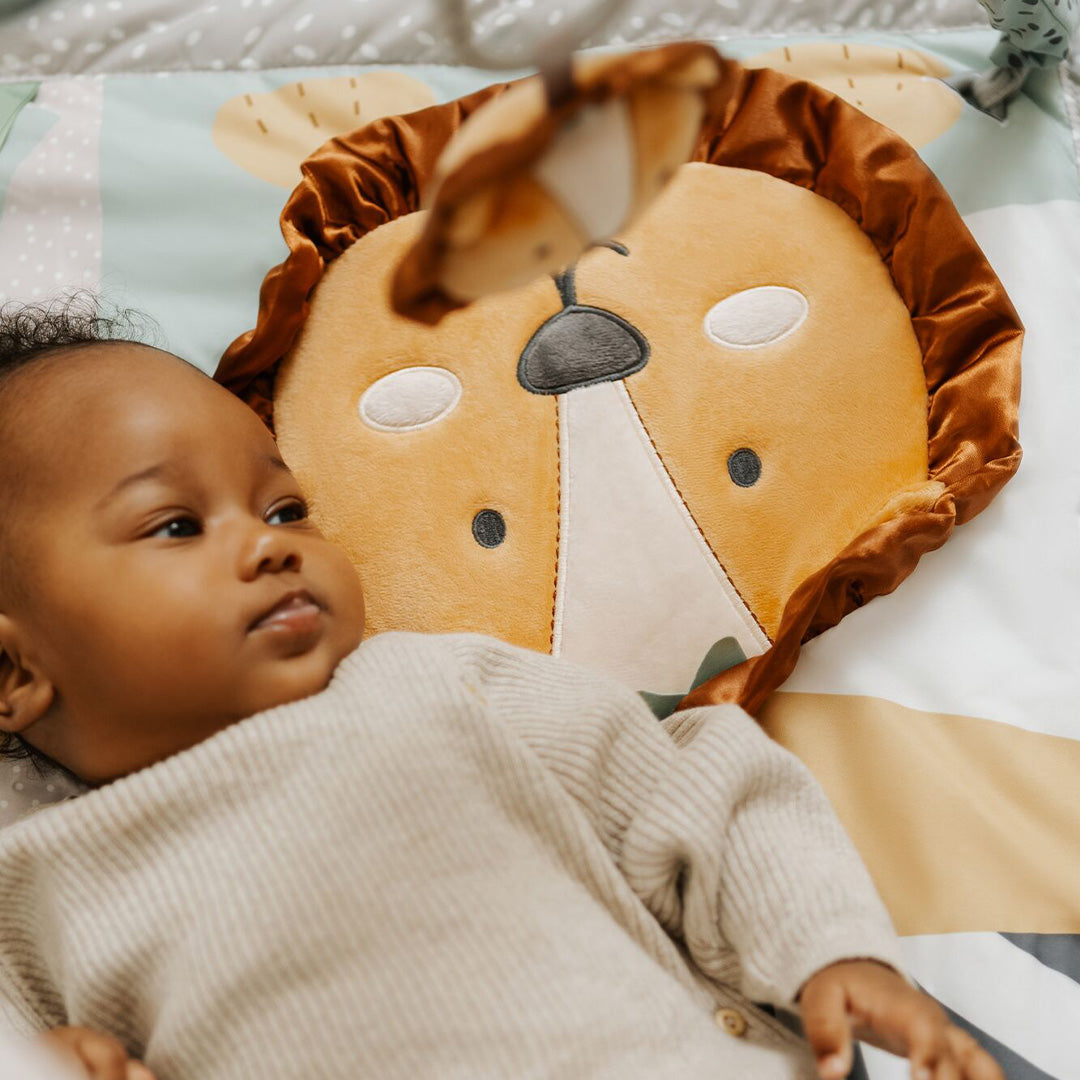 A baby rests on the Nuby Animal Adventures Baby Play Mat with Gym, lying beside a plush lion face pillow and looking up at a lion-shaped mobile and sensory toys overhead.