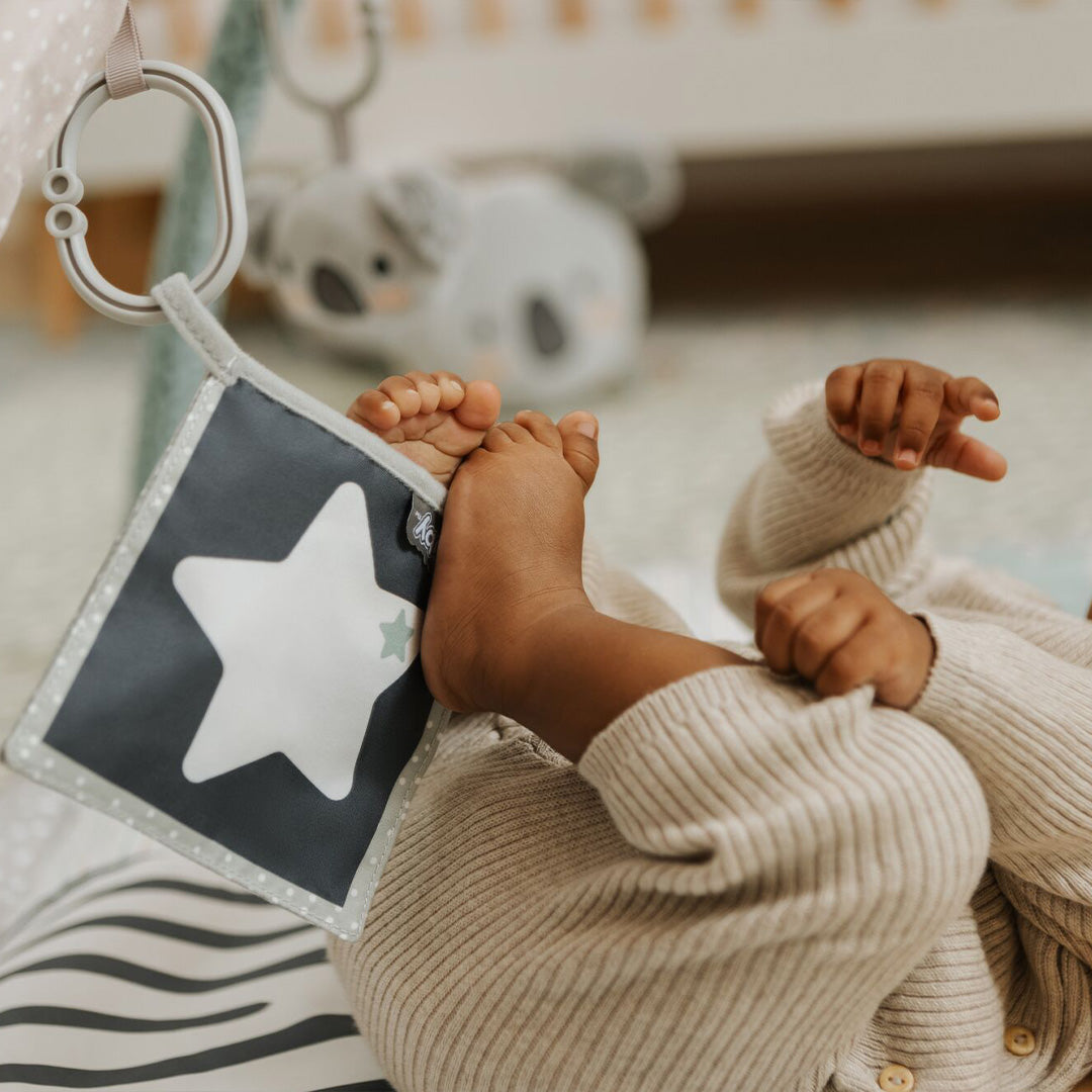 A baby in a ribbed beige outfit lies on the Nuby Animal Adventures Baby Play Mat with Gym, playing with their feet. A soft sensory toy with a white star hangs from the gym by a plastic ring, and a stuffed animal is in the background.