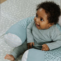 A smiling baby in a ribbed, light gray outfit sits supported by the Nuby Animal Adventures Baby Sit Me Up Floor Seat, which features blue and white patterns, on a soft dotted blanket.