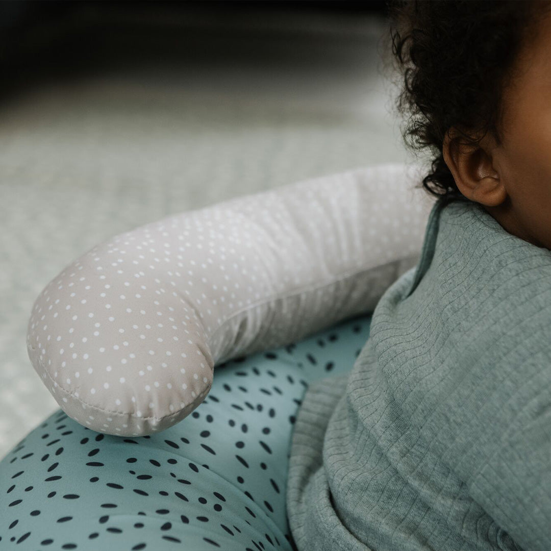 A young child in green sits in the Nuby Animal Adventures Baby Sit Me Up Floor Seat, featuring a beige polka-dotted pillow and blue patterned cushion; only part of the child’s face and upper body is visible.