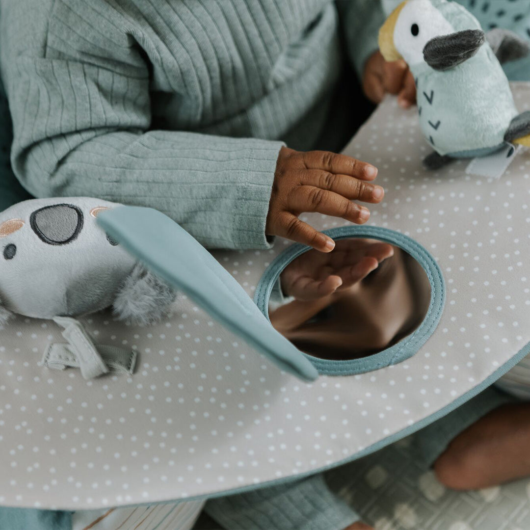 A young child in a green ribbed outfit plays on a soft, polka-dotted mat with a plush parrot and touches a round mirror beside the Nuby Animal Adventures Baby Sit Me Up Floor Seat with activity tray.