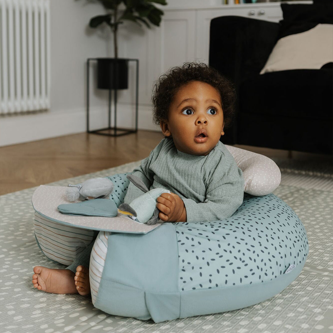 A baby with curly hair sits in a soft, blue Animal Adventures Baby Sit Me Up Floor Seat by Nuby on a play mat, looking up in surprise while holding a fabric toy. A plant and furniture are visible in the background.