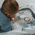 A baby in a blue sweater lies on their stomach, exploring the Nuby Animal Adventures Tummy Time Water Mat featuring lion, bear, and sheep illustrations on a soft, patterned surface.
