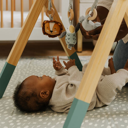 A baby lies on a patterned mat under the Nuby UK Animal Adventures Nursery Bundle wooden play gym, reaching for animal toys as an adult adjusts one. A crib, also from newborn nursery essentials, appears in the background.