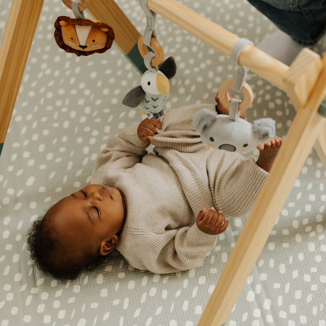 A baby in a beige outfit lies on a patterned mat, reaching up to play with plush animal toys hanging from the Nuby Animal Adventures Wooden Play Gym, enjoying sensory play and gentle movement.