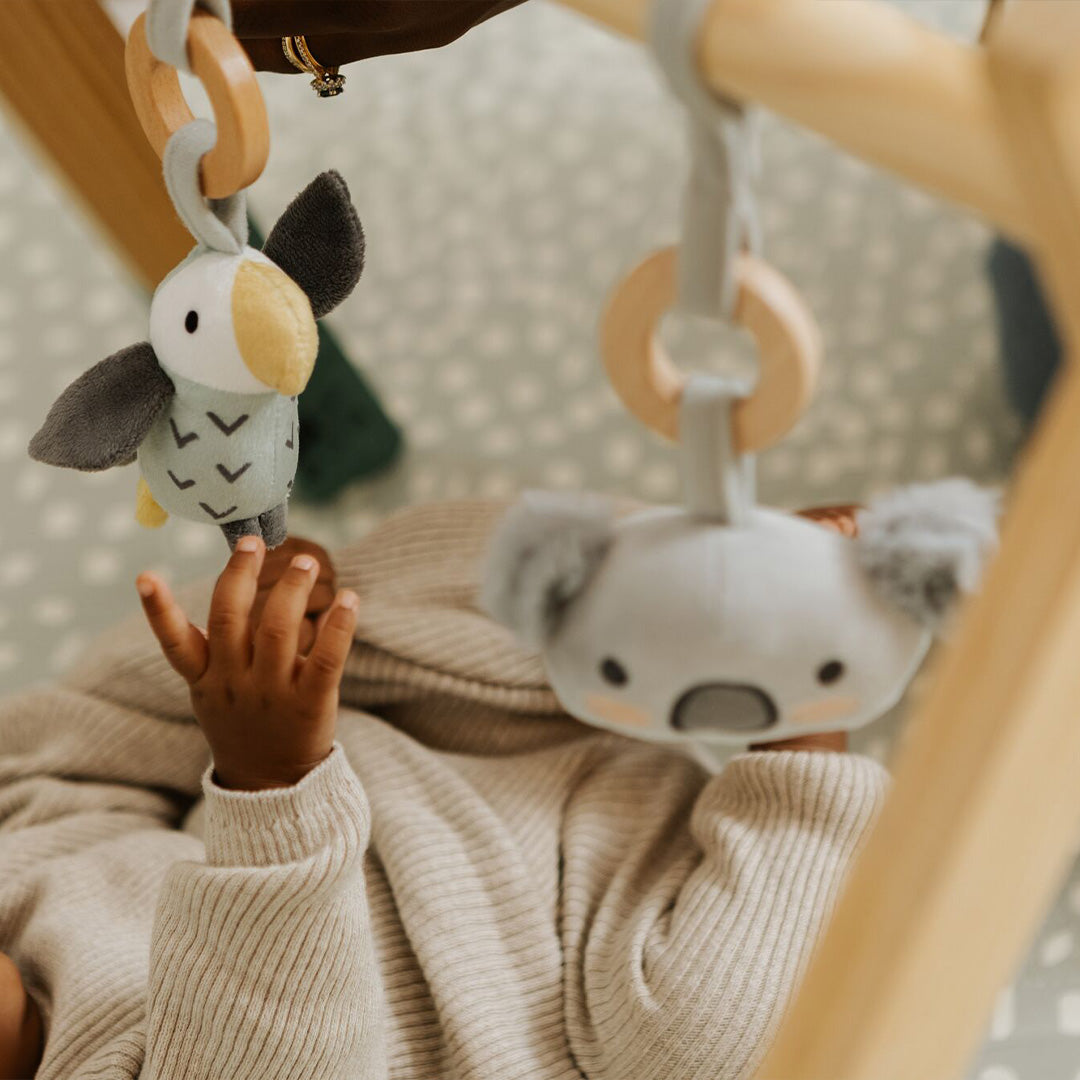 A baby in a beige outfit reaches for plush koala and bird toys hanging from the Nuby Animal Adventures Wooden Play Gym, while the dotted, light-colored play mat underneath offers added comfort for sensory play.