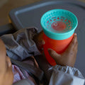 The all around beaker by Nuby in a red and aqua colourway. Being held in the baby's hands.