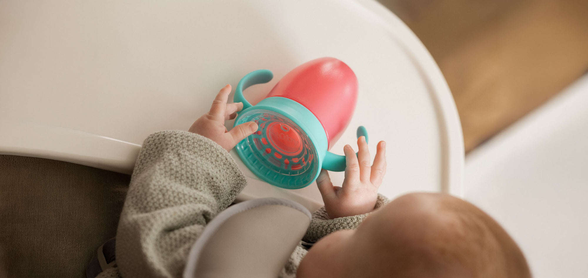 A baby holding a cup from Nuby that has a turquoise lid and handles with a red cup lower section. The baby is sat at a high chair with a small plastic table to rest their arms and the cup onto.