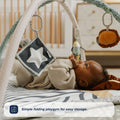 A baby in beige reaches for hanging toys on the Nuby UK Animal Adventures Playtime Set, a patterned play mat with simple folding playgym for easy storage. A crib appears in the background.