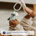 A baby in a ribbed beige outfit lies on the Nuby UK Animal Adventures Playtime Set, reaching for a soft hanging parrot toy. Text overlay: Removable hanging toys for babys sensory play time.