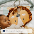 A baby relaxes on the Nuby UK Animal Adventures Playtime Set, a soft mat with a lion face design, gazing at a plush toy overhead. The set offers comfort and gentle textures for sensory exploration during tummy time.