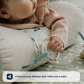 A baby in a pink sweater rests on a patterned pillow, holding the Nuby UK Animal Adventures Playtime Set ring toy. Text below reads: Three sensory features that rattle and crinkle—perfect for tummy time roller fun.