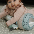 A baby in a pink sweater relaxes on the Nuby UK Animal Adventures Playtime Set, hugging a tummy time roller with a mirror and polka dots, and holding a plush bird.