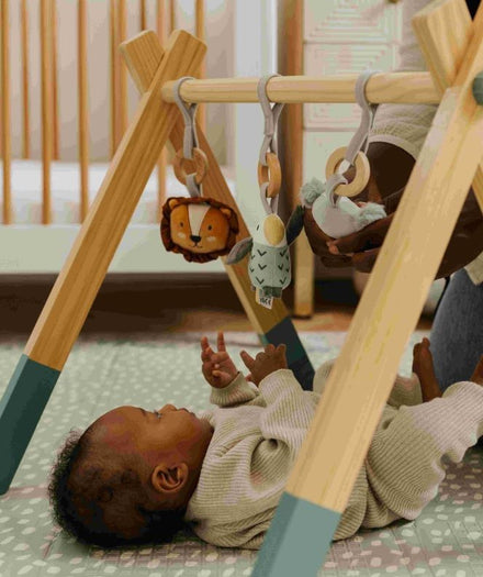 A baby lying down and playing with their Animal Adventures Wooden Play Gym.