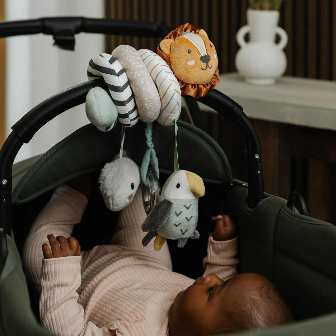 A baby in a pale pink outfit lies in a stroller, reaching for the Animal Adventures Pram Spiral Activity Toy with plush lion, bird, and whale hanging from the handle. A white vase is visible on a table in the background.