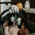 A baby in a pale pink outfit lies in a stroller, reaching for the Animal Adventures Pram Spiral Activity Toy with plush lion, bird, and whale hanging from the handle. A white vase is visible on a table in the background.